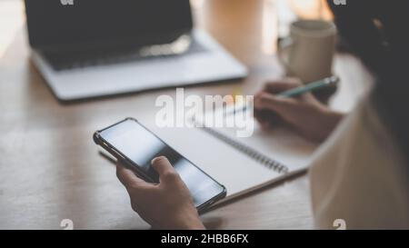 Eine junge Frau nutzt ein Smartphone und schreibt Informationen in ein Notebook. Stockfoto