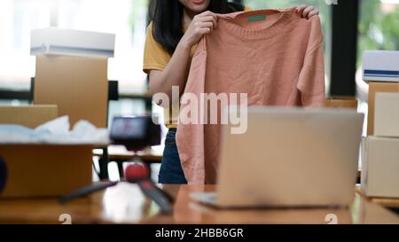 Frauen zeigen den Verkauf von Hemden online von zu Hause aus. Sie nutzt ihren Laptop, um über soziale Medien zu verkaufen. Online-Shopping-Konzepte. Stockfoto