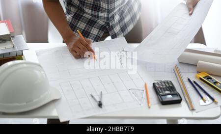 Designer-Mann mit einem Bleistift, der Hauspläne, Schutzhelm und Modellhäuser mit Zeichenwerkzeugen auf einem Schreibtisch untersucht, Home Design Architect. Stockfoto