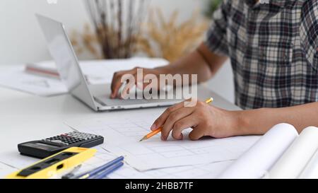 Der Mensch schreibt mit einem Bleistift auf den Hausplan und legt seine Hand auf die Laptop-Tastatur, den Hausplan mit Zeichenwerkzeugen auf dem Schreibtisch, Architektendesign Stockfoto