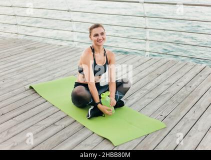 Junge glückliche Sportlerin in Sportbekleidung, die auf der Holzterrasse am Strand auf der Matte sitzt. Gesundes Lifestyle-Konzept Stockfoto