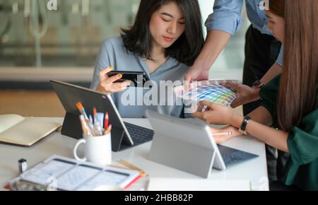 Das Grafikdesigner-Team sucht nach einer Farbkarte, um ein Projekt mit einem Laptop und einem Tablet auf dem Schreibtisch zu entwerfen. Stockfoto