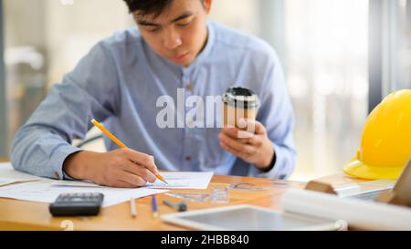 Der junge Designer, der einen Kaffeebecher zum Mitnehmen in der Hand hat, verwendet einen Bleistift, um ein Hausdesign zu entwerfen. Stockfoto