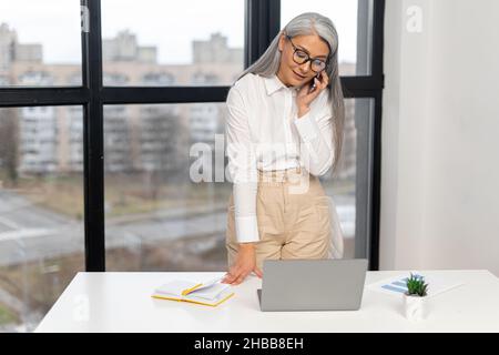 Seriöse alte Geschäftsfrau mit elegantem Casual Shirt und stylischer Brille, die im modernen Bürobereich über das Smartphone steht und chattet. Reife herrische Dame, ältere zielstrebige Führerin Konzept Stockfoto