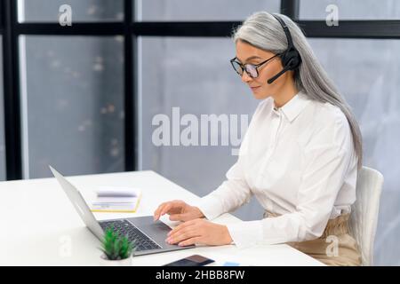 Eine ältere, grauhaarige Geschäftsfrau im Headset sitzt am Schreibtisch, trifft sich online mit dem Kunden, Videoanrufe über den Laptop vom Büro aus. Ältere Frau, die auf den Bildschirm schaut Stockfoto