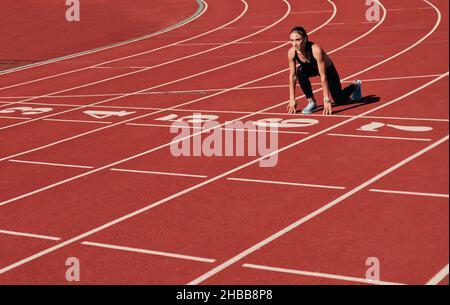 Junge Läuferin Frau in Sportbekleidung, die sich bereit für den Sprint bei niedrigem Start auf der Stadionstrecke mit roter Beschichtung an einem hellen sonnigen Tag gemacht hat Stockfoto