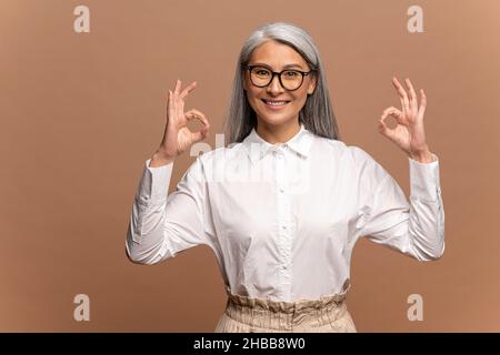 Porträt einer zufriedenen asiatischen älteren Frau mit grauen Haaren in formeller weißer Bluse stehend, Blick auf die Kamera mit OK Zeichen Geste, Innenaufnahme Studio isoliert auf beigem Hintergrund Stockfoto