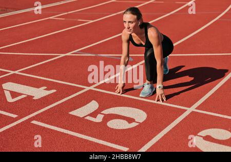 Junge Läuferin Frau in Sportbekleidung, die sich bereit für den Sprint bei niedrigem Start auf der Stadionstrecke mit roter Beschichtung an einem hellen sonnigen Tag gemacht hat Stockfoto