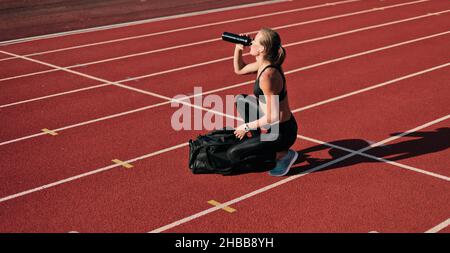 Junge Sportlerin in Sportbekleidung bereitet sich auf das Training vor, während sie an sonnigen Tagen die Sporttasche öffnet und Wasser auf der Strecke des Stadions trinkt Stockfoto