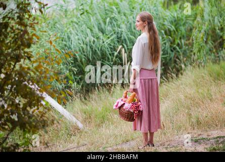 Junge lächelnde Frau in Vintage Retro-Stil Kleidung hält einen Picknickkorb in der Hand vor dem Hintergrund des Schilf auf dem See Stockfoto