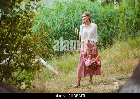 Junge lächelnde Frau in Vintage Retro-Stil Kleidung hält einen Picknickkorb in der Hand vor dem Hintergrund des Schilf auf dem See Stockfoto