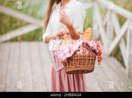 Junge blonde Frau mit langen Haaren in Vintage-Kleidung im Retro-Stil steht mit einem Picknickkorb auf einem hölzernen Pier des Sees Stockfoto