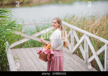 Junge blonde Frau mit langen Haaren in Vintage-Kleidung im Retro-Stil steht mit einem Picknickkorb auf einem hölzernen Pier des Sees Stockfoto