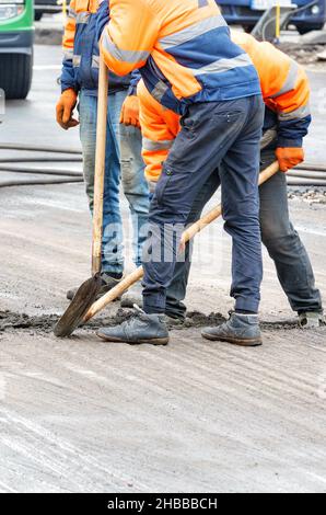 Straßenarbeiter in orangefarbenen Overalls verwenden Schaufeln, um Rückstände von der alten Straßenoberfläche zu entfernen. Stockfoto