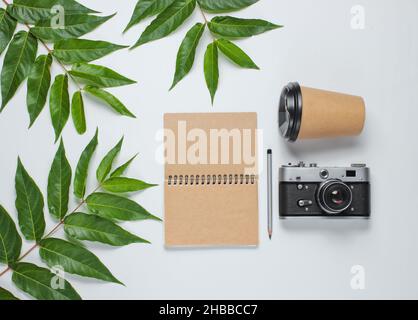 Bastelnotizbuch mit Bleistift, Kaffeetasse, Retro-Kamera auf weißem Hintergrund mit grünen Blättern. Das Konzept der Einheit mit der Natur. Draufsicht Stockfoto