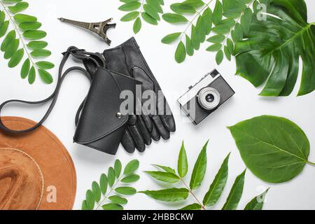 Jorney nach Paris. Damenzubehör (Hut, Handschuhe, Tasche), Retro-Kamera, Figur des Eiffelturms auf weißem Hintergrund mit grünen Blättern. Draufsicht Stockfoto