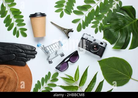 Jorney nach paris. Damenzubehör (Hut, Handschuhe, Sonnenbrille), Retro-Kamera, Figur des Eiffelturms auf weißem Hintergrund mit grünen Blättern. Oben Stockfoto