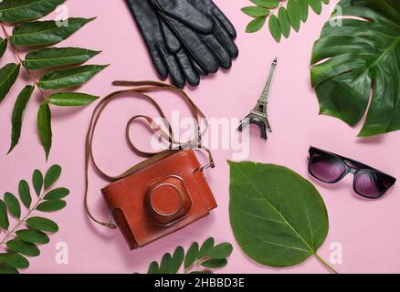 Jorney nach Paris. Damenzubehör, Retro-Kamera, Figur des Eiffelturms auf rosa pastellfarbenem Hintergrund mit grünen Blättern. Draufsicht Stockfoto