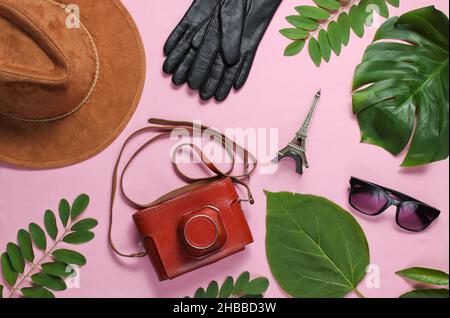 Jorney nach Paris. Damenzubehör, Retro-Kamera, Figur des Eiffelturms auf rosa pastellfarbenem Hintergrund mit grünen Blättern. Draufsicht Stockfoto