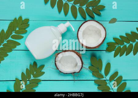 Minimalistische Schönheit Stillleben. Zwei Hälften gehackte Kokosnuss und weiße Flasche Creme mit grünen Blättern auf blauem Holzhintergrund. Kreative Mode Stockfoto