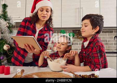 Fröhliche fröhliche Familie in karierten Kleidern, schöne junge liebevolle Mutter in Weihnachtsmütze und ihre beiden entzückenden Kinder, Junge und Mädchen Vorbereitung toge gekleidet Stockfoto