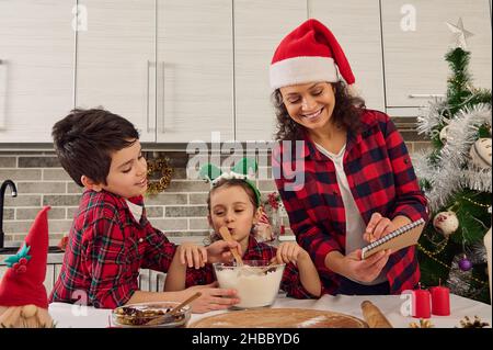 Fröhliche fröhliche Familie in karierten Kleidern, schöne junge liebevolle Mutter in Weihnachtsmütze und zwei entzückende Kinder, Junge und Mädchen, die sich zusammen vorbereiten Stockfoto