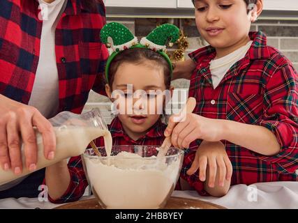 Das hübsche kleine Mädchen sitzt zwischen ihrer liebenden Mutter, die etwas Milch in eine Schüssel mit Mehl gießt, und ihrem fröhlichen älteren Bruder, der trocken mischt und knetet Stockfoto