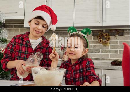 Fröhliche Bruder und Schwester, süße kaukasische Kinder verbringen Zeit zusammen in der Küche zu Hause. Liebenswert Junge gießt Milch und Baby Mädchen knetet Teig für Chris Stockfoto
