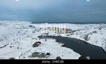 Luftaufnahme eines langen Flusses, der in der Wintersaison durch das kleine Dorf fließt, umgeben von schneebedeckten Feldern. Fliegen über weißen Wiesen, Gebäuden und Stockfoto