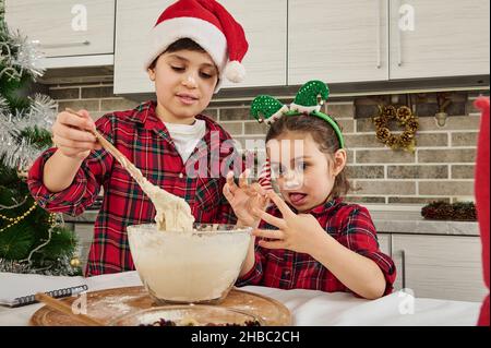 Entzückende europäische Kinder, die zusammen Spaß haben und Teig für Weihnachtskekse, Lebkuchen, Muffins in der heimischen Küche zubereiten. Bruder in Santa hat Stockfoto