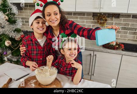 Glückliche Mutter in Santa hat hält ein Handy hone in ausgestreckten Händen und lächelt machen Selfie umarmt ihren schönen Sohn und Tochter, Vorbereitung zusammen Stockfoto