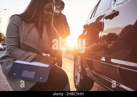 Versicherungsvertreter untersucht das Auto, während der Fahrer auf dem Mobiltelefon spricht. Unfallanspruchsprozess. Afroamerikanische Frau, die den Schaden an der überprüft Stockfoto