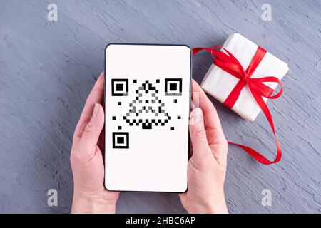 Hände, die ein Smartphone, Tablet, Mobiltelefon mit einem qr-Code-Bildschirm in Form eines Weihnachtsbaums und einer Geschenkbox auf grauem Hintergrund halten. Weihnachten Stockfoto