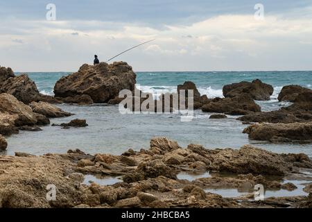 Ein eingeflügelter Fischer, der an einem stürmischen Wintertag an einem felsigen Ufer fischt. Stockfoto