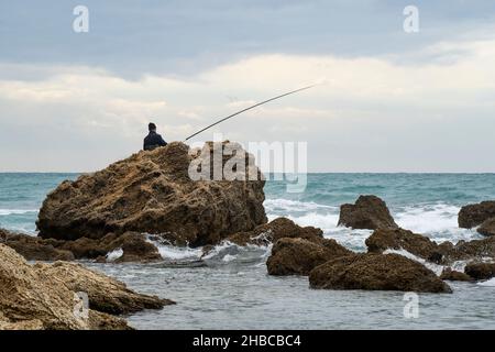 Ein eingeflügelter Fischer, der an einem stürmischen Wintertag an einem felsigen Ufer fischt. Stockfoto