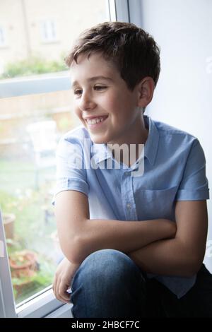 Ein Porträt eines lächelnden Jungen, während er in seinem Haus auf einer Fensterbank saß Stockfoto
