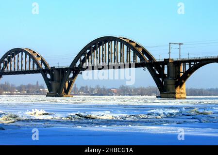 Gewölbte Eisenbahnbrücke über den Fluss im Winter. Eine schöne alte Brücke, der Fluss Dnjepr ist mit Eis und Schnee bedeckt. Dnipro Stadt, Dnepropetrovsk Stockfoto