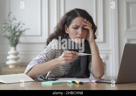 Ungesunde spanierin in warmen Strickschal gewickelt und ängstlich auf Digitalthermometer schauend Stockfoto