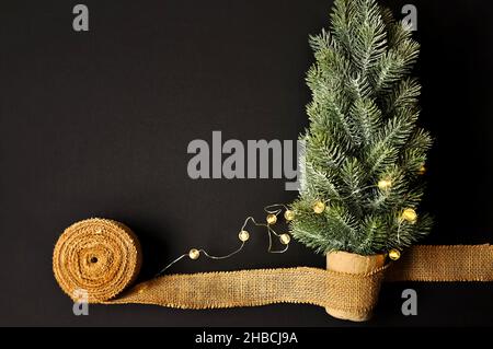 Vorbereitung von Dekorationen für Winterfeste. Künstlicher Weihnachtsbaum, natürlicher Jutefaserstreifen, dunkler Hintergrund, gemütliche Beleuchtung Stockfoto