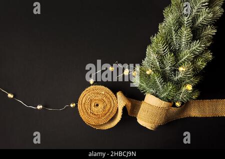 Vorbereitung von Dekorationen für Winterfeste. Künstlicher Weihnachtsbaum, natürlicher Jutefaserstreifen, dunkler Hintergrund, gemütliche Beleuchtung Stockfoto