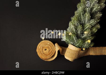 Vorbereitung von Dekorationen für Winterfeste. Künstlicher Weihnachtsbaum, natürlicher Jutefaserstreifen, dunkler Hintergrund, gemütliche Beleuchtung Stockfoto