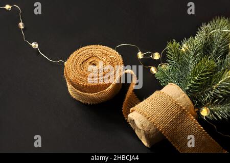 Vorbereitung von Dekorationen für Winterfeste. Künstlicher Weihnachtsbaum, natürlicher Jutefaserstreifen, dunkler Hintergrund, gemütliche Beleuchtung Stockfoto