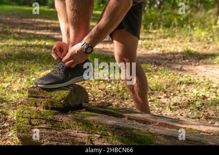 Der Sportler bindet die Schnürsenkel seiner Beine in Turnschuhen aus der Nähe der Sportler läuft im Park draußen, um den Wald, Eichen grünen Gras youn Stockfoto