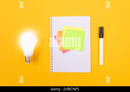 Glühende Energiesparlampe und Notizblatt mit klebriger Notiz auf gelbem Tisch, leere Schablone mit Kopierraum - Draufsicht, flaches Lay Stockfoto