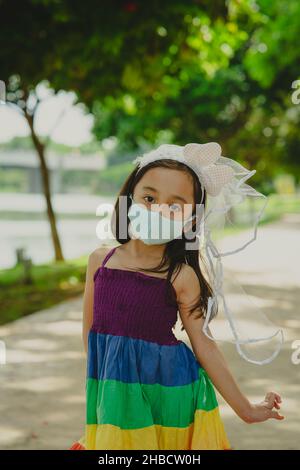 Kleines Mädchen trägt Gesichtsmaske mit Regenbogenkleid genießen im Freien im Park. Neuer Normalwert. Stockfoto