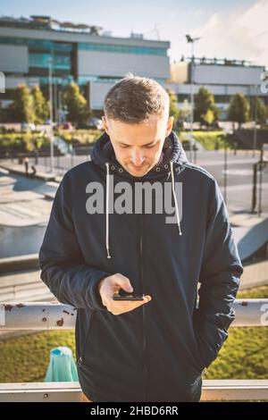 Lifestyle, Freizeit, Telekommunikationskonzept. Mann mit dunkler Jacke, der während eines sonnigen Tages im Stadthintergrund während eines sonnigen und warmen Tages das Handy benutzt Stockfoto