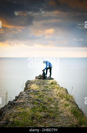 Rüde kuschelt Hund in der Natur vor dem Meer mit dramatischem Himmel im Hintergrund. Konzept der Freundschaft zwischen Tieren und Menschen. Stockfoto