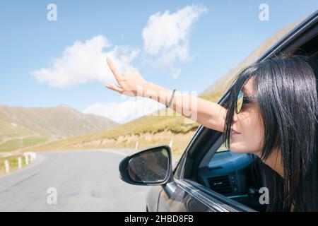 Weibliche Kaukasierin Frau aus Autofenster zeigt Finger nach links mit Asphalt Straße Hintergrund. Copypaste-Reisekonzept. Stockfoto