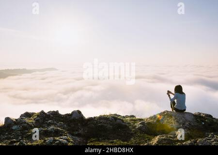 Brünette Frau sitzt auf der Klippe hält Smartphone und macht ein Foto des Sonnenaufgangs über den Wolken. Konzept von Reisefreude und Erinnerungen. Stockfoto
