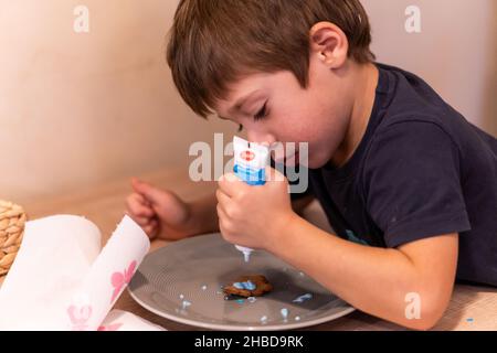 POZNAN, POLEN - 14. Nov 2021: Ein kleiner Junge schmückt Ingwerkekse auf einem grauen Teller auf einem Tisch in Poznan, Polen Stockfoto